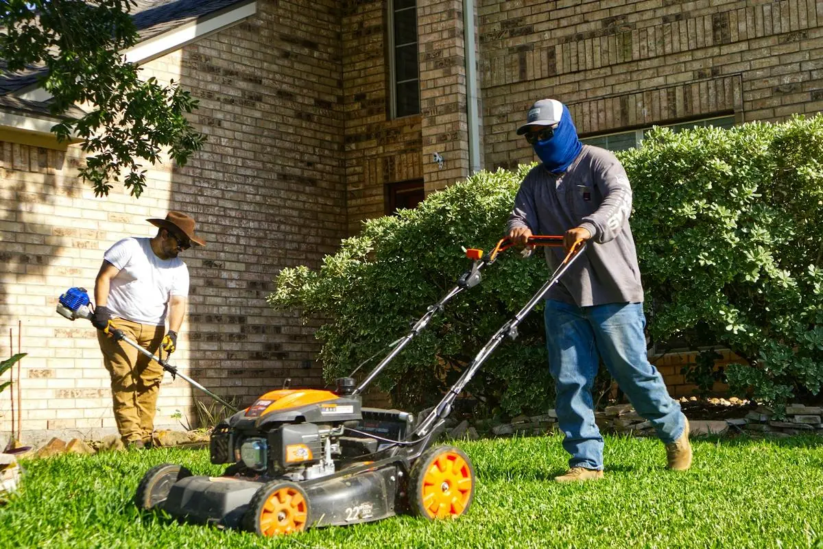 Two crew members mowing and string-trimming the front lawn of a brick house